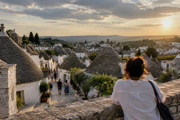 An evening view across trulli rooftops in Alberobello, a person leaning on a low stone wall in warm light