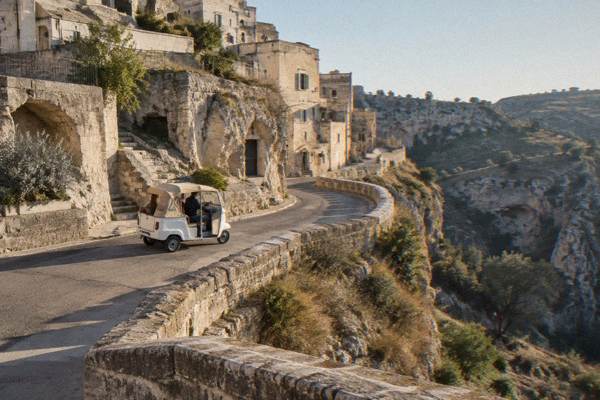 A three-wheeled tuk-tuk on a narrow road along the ravine edge below pale cave houses in Matera