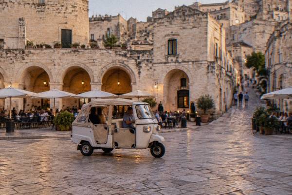 A three-wheeled tuk-tuk crossing an evening square of pale stone arches and cafe tables in Matera