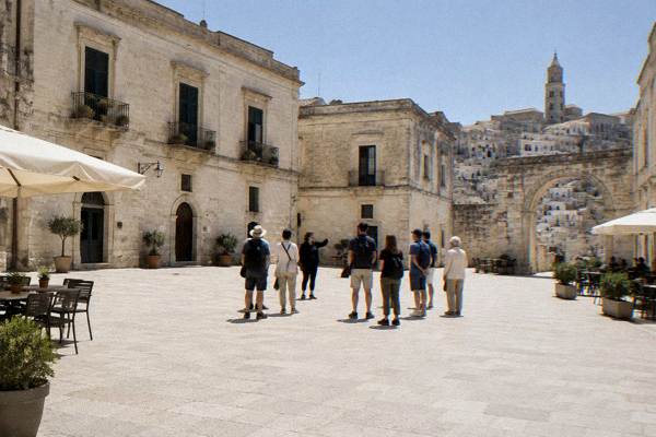 A small group of walkers gathered in a broad pale-paved square in Matera, cream stone buildings all around