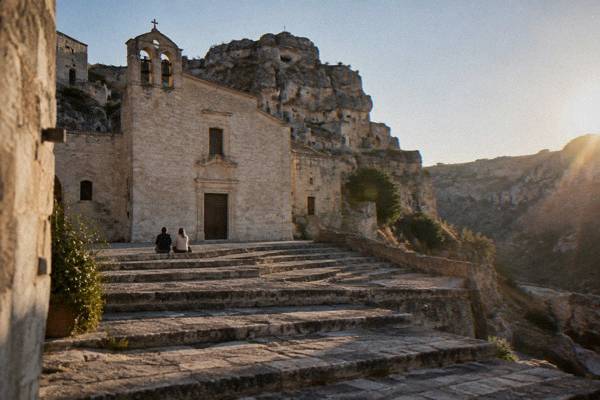 A wide stepped stone square in front of a pale stone church with a bell gable in Matera, two people sitting on the steps