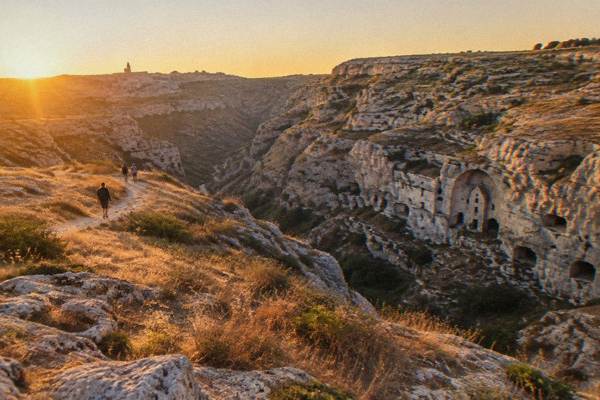 Walkers on a path across the pale limestone plateau at golden hour near Matera, canyon cliffs with rock-cut openings