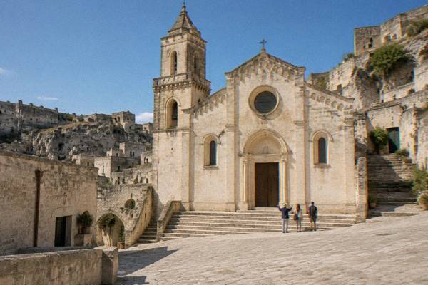 A guide and two visitors near a pale stone church on a small sloping square among cave houses in Matera