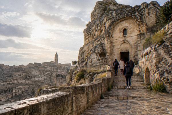 A worn stone path climbing to a rock-cut church set into a limestone pinnacle above old Matera, visitors from behind