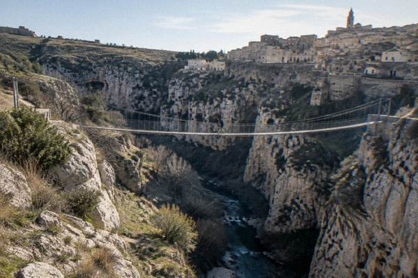A narrow suspension footbridge over a deep rocky canyon near Matera, one walker tiny on the bridge