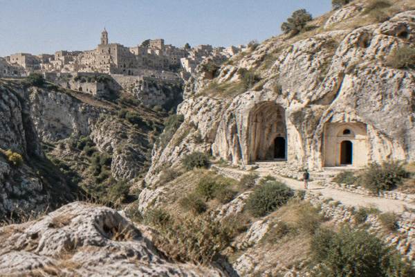 Rock-cut church entrances in a pale limestone slope across the canyon from the old town of Matera, a tiny visitor on the path