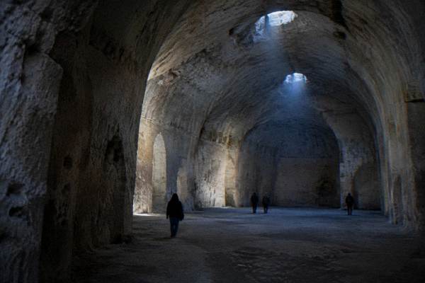 A vast underground stone chamber with arched pale walls and a few small visitors in Matera