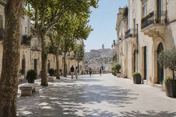 A pedestrian street of cream stone buildings and plane trees in Matera's upper town, walkers heading toward the old districts