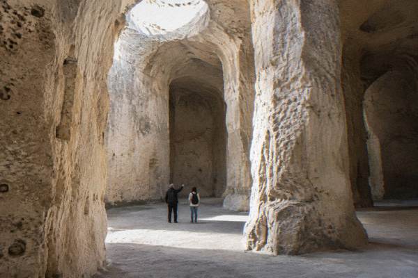 A guide and a visitor small in a huge excavated tufa quarry chamber with tall pale walls in Matera
