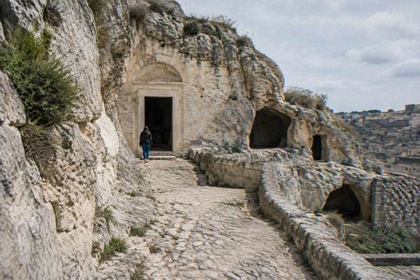 A modest rock-hewn church doorway cut into a limestone slope in Matera, with a worn stone path leading to it