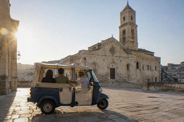 A three-wheeled tuk-tuk stopped on the paved forecourt of a pale-stone cathedral in Matera