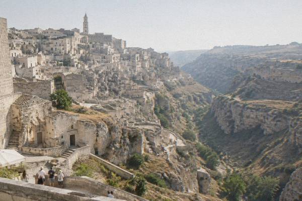 A broad view of stacked pale cave dwellings across the ravine in Matera, a tiny group of visitors on a terrace below
