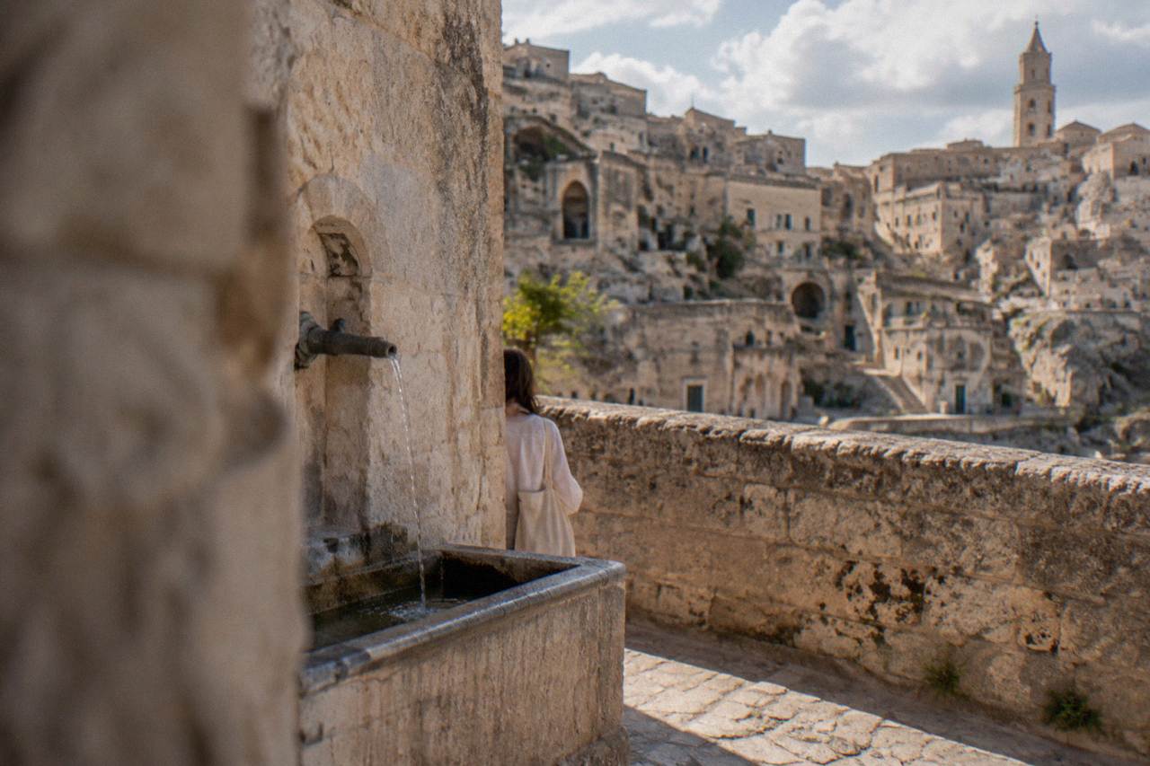 A stone drinking fountain set into a limestone wall in Matera, with cave dwellings in the background.