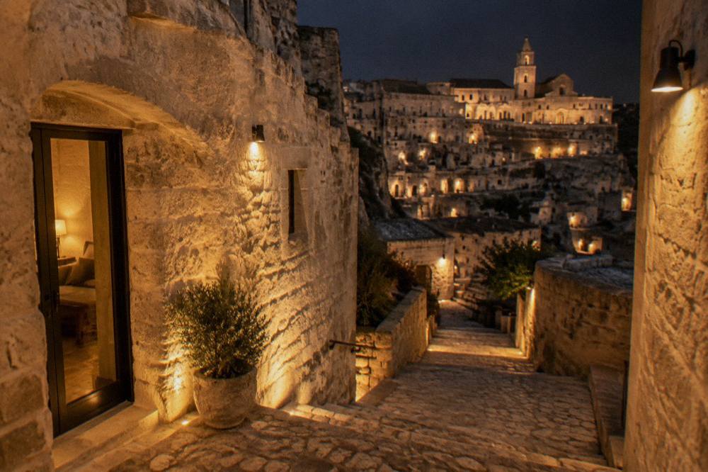 A narrow stepped lane in the Sassi of Matera at night, stone walls lit by warm lamps, with a cave hotel entrance on the left