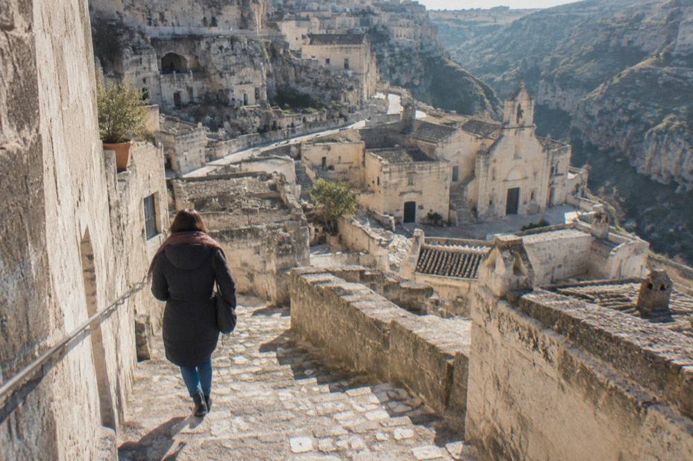 A visitor in a warm coat and scarf walking down a worn stone staircase in the Sassi of Matera on a winter morning