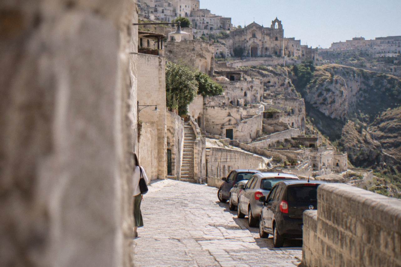 A narrow stone street in the Sassi of Matera with pale cave dwellings rising in terraces above parked cars