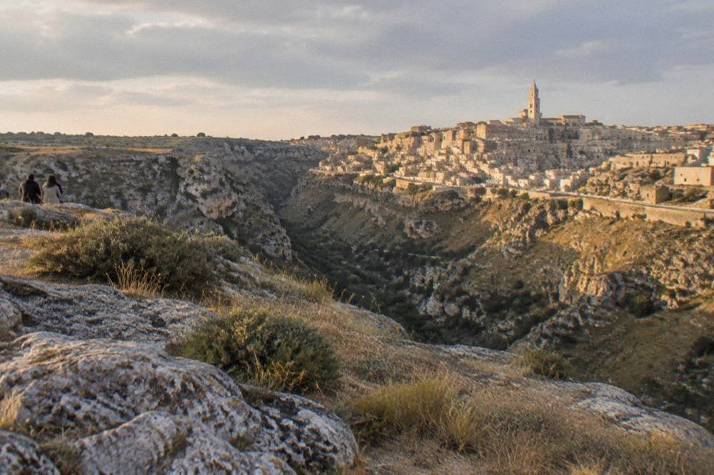 A hillside city of pale stone buildings and cave dwellings seen from across a deep ravine at golden hour