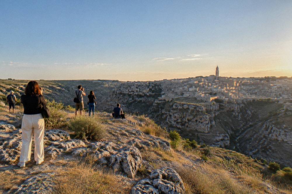 A small crowd of people with cameras standing on a rocky ledge at dusk facing a glowing stone city