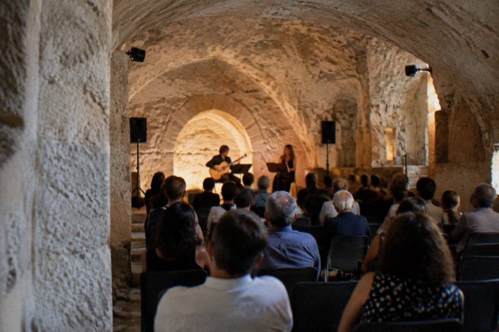 An audience seated in a stone-vaulted underground hall before a small stage