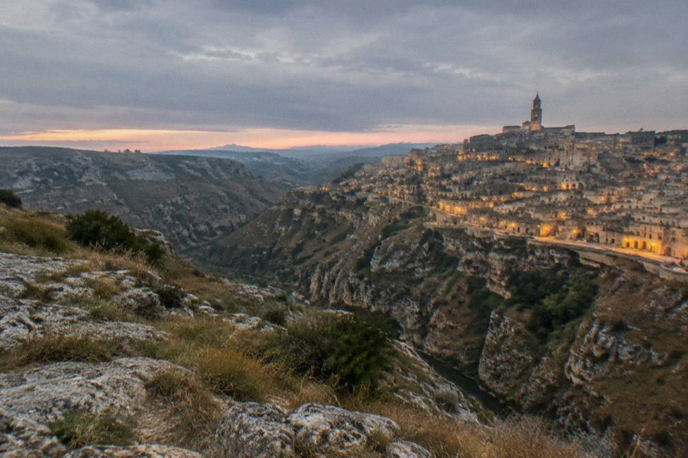 A panoramic view of the Sassi districts across the Gravina canyon at dusk with lit windows in the cave facades
