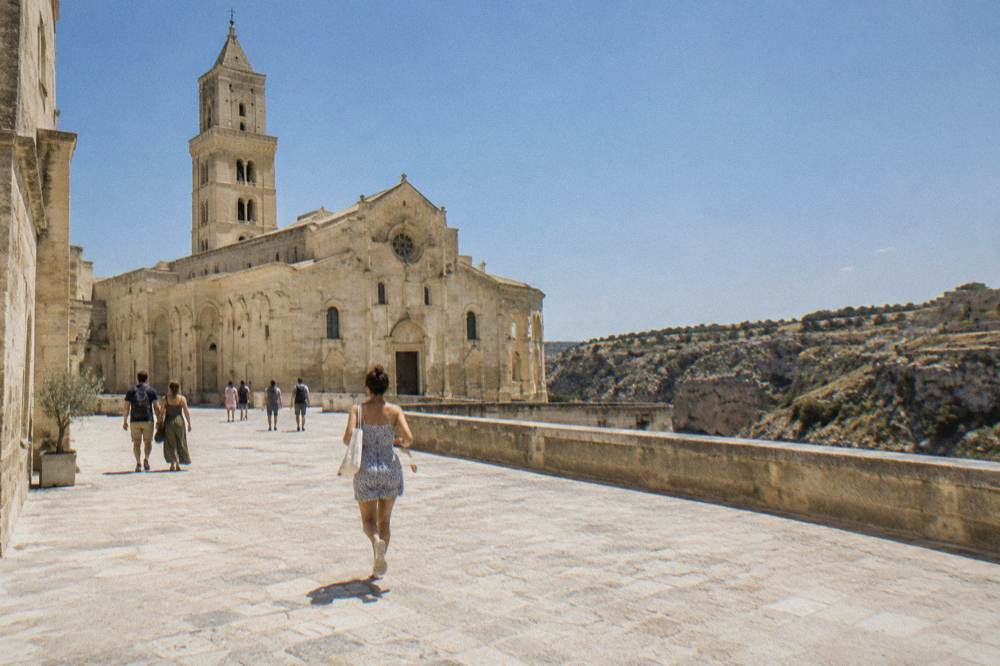 Visitors walking on the stone terrace in front of Matera Cathedral with the canyon landscape beyond