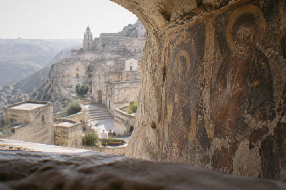 Frescoed rock wall inside a Matera cave church, painted figures darkened by age