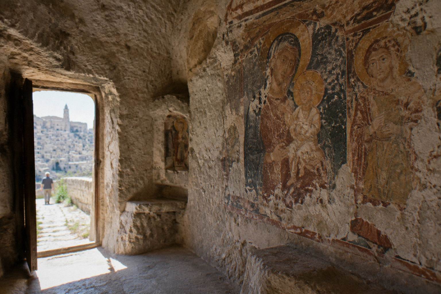Weathered fresco on the wall of a small rock-cut church, lit by a narrow doorway