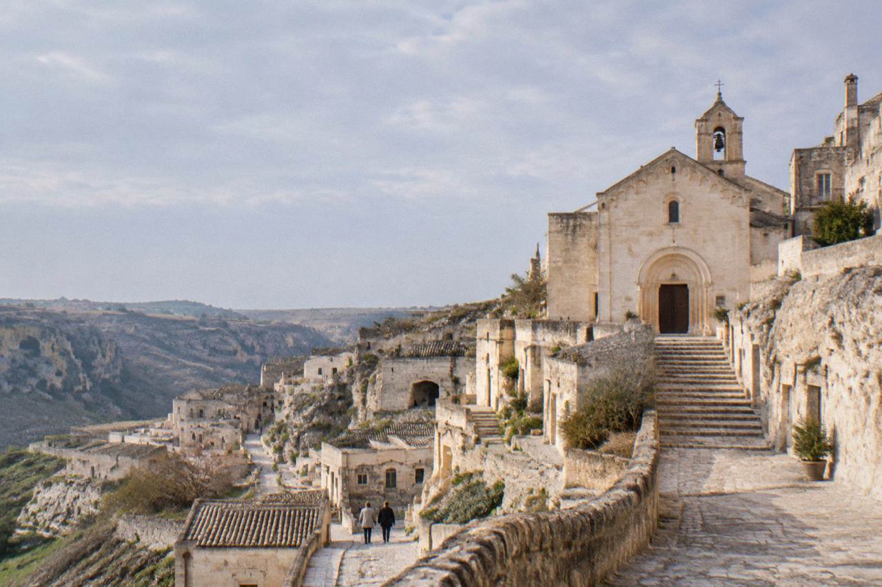 A pale stone church facade with a simple arched portal rising above narrow stepped lanes carved into the tufa hillside of Matera