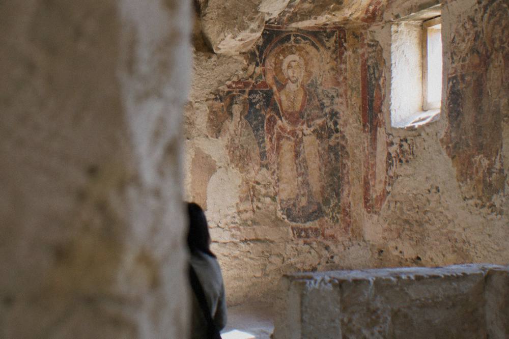 Interior of a rock-cut church in Matera with faded frescoes on the carved stone walls lit by natural light from a small window