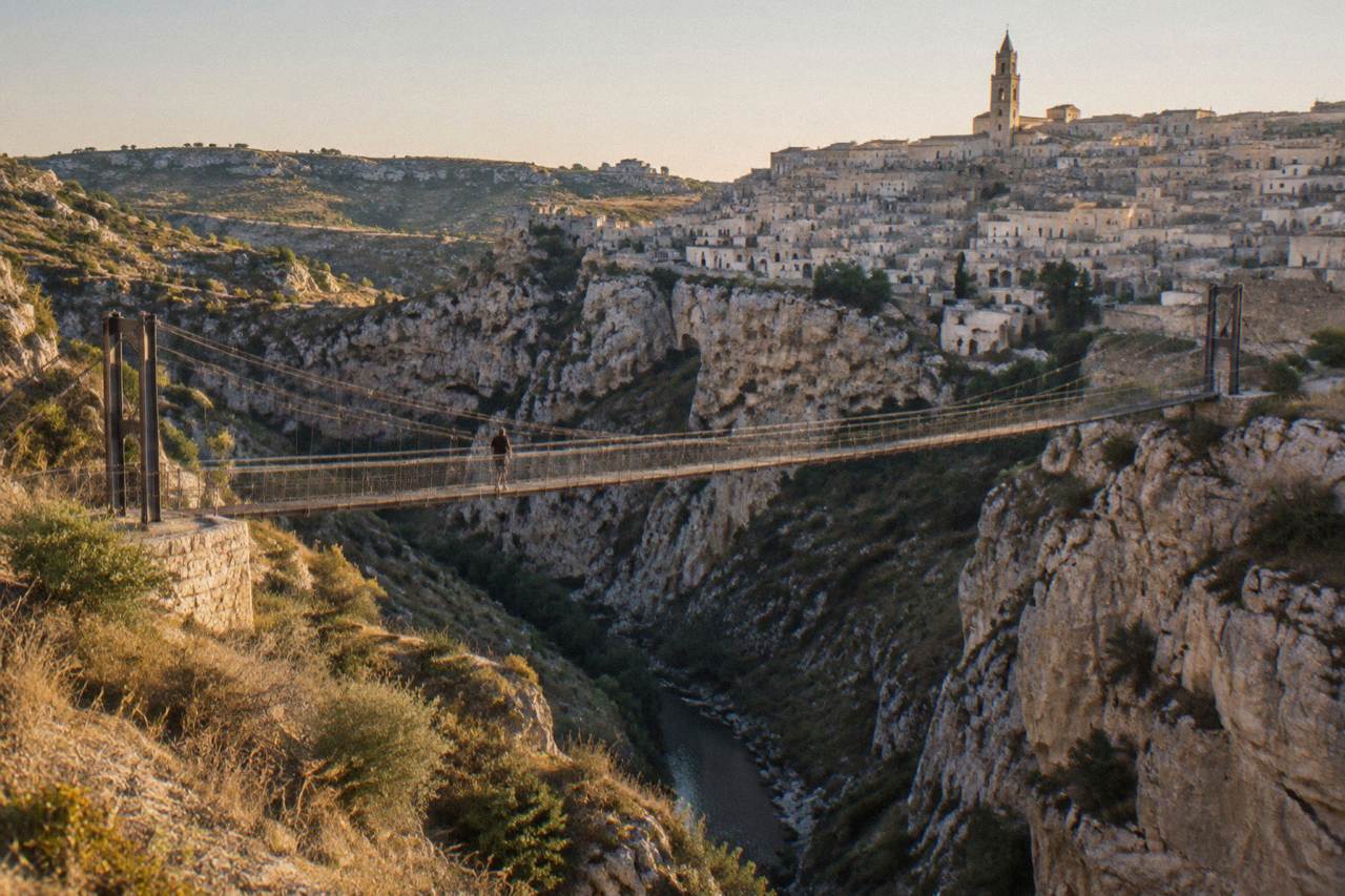 A suspension bridge spanning a deep limestone canyon at dusk, with a cave town visible on the far side