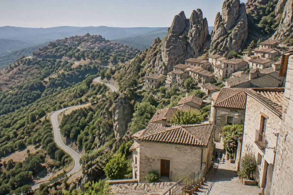 A narrow mountain road curving through wooded slopes toward a ridge-top village