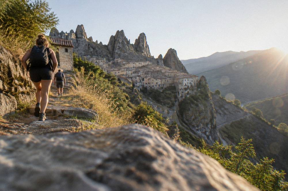 Hikers on a rocky path with a stone village visible on a cliff in the distance