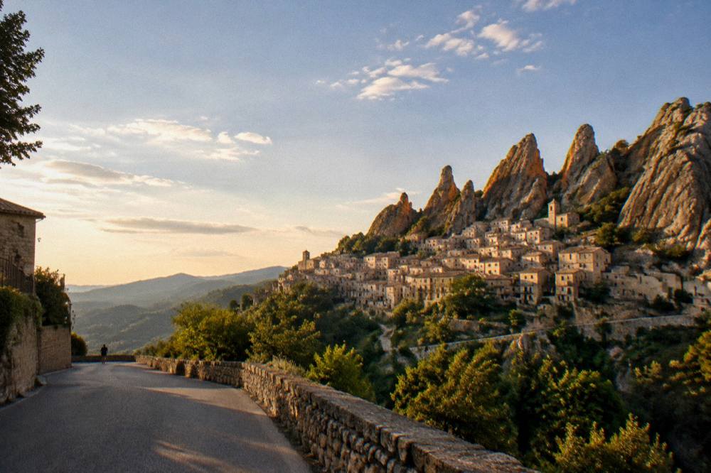 A stone village silhouette on a ridge under a wide evening sky seen from a valley road