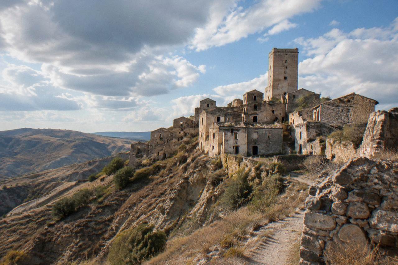 An abandoned hilltop village of empty stone houses and a tall bell tower rising above bare clay hills under a wide sky