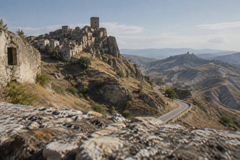 A winding rural road through bare clay badlands in southern Italy with a distant abandoned village on a ridge