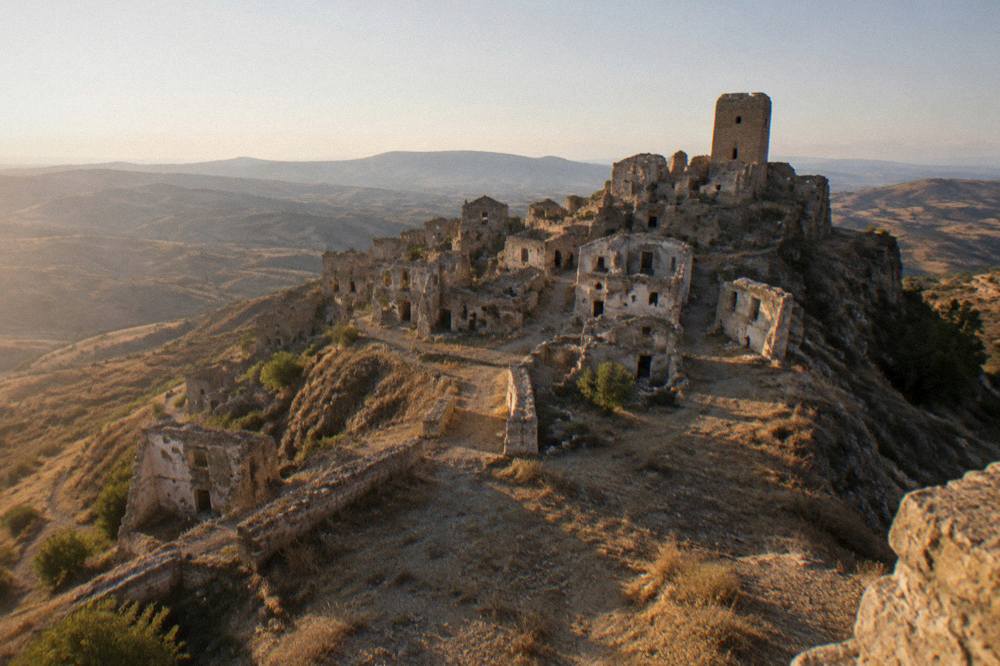 A wide view of an abandoned hill town silhouetted against a pale sky at dusk, with empty window openings visible on ruined facades