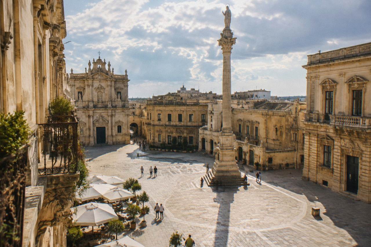 A sunlit baroque piazza in Lecce with pale stone facades and a tall column in the centre