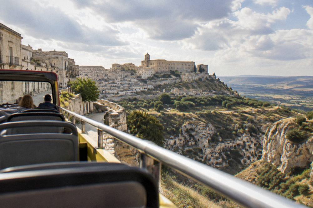 An open-top sightseeing bus parked on a street above a canyon of pale cave dwellings