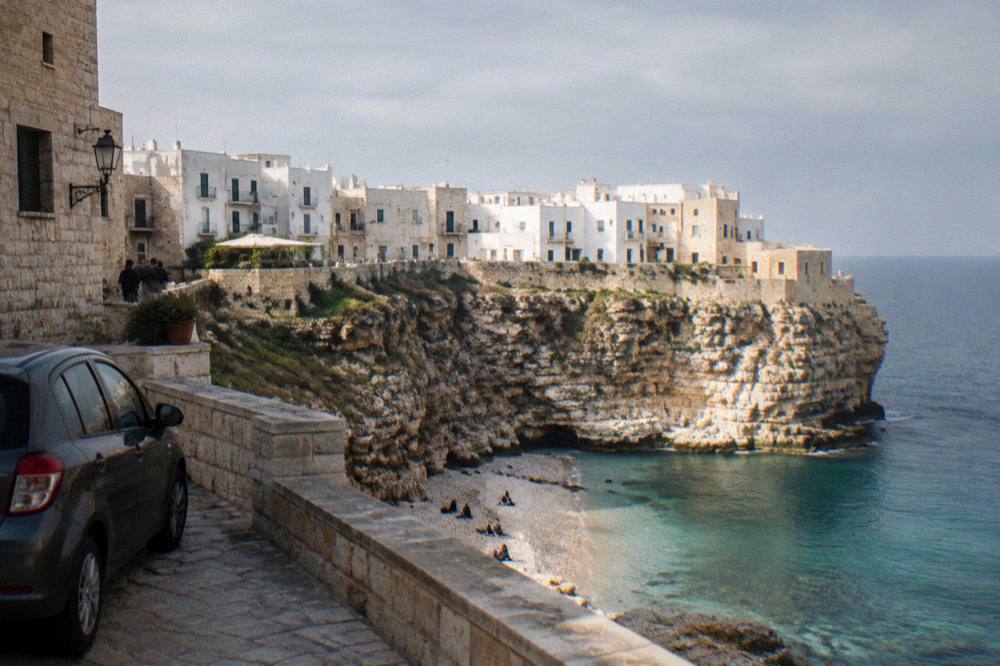 A parked car and stone buildings at the edge of a coastal piazza in late light