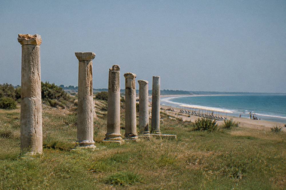 A wide sandy Ionian shoreline with shallow water and a line of umbrellas under a summer sky
