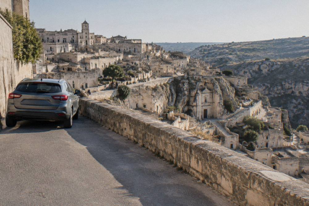 A rental car parked on a wide road above the Sassi district of Matera at midday