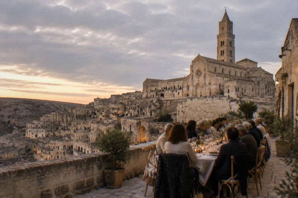 A long table of diners on a stone terrace overlooking cave houses at dusk