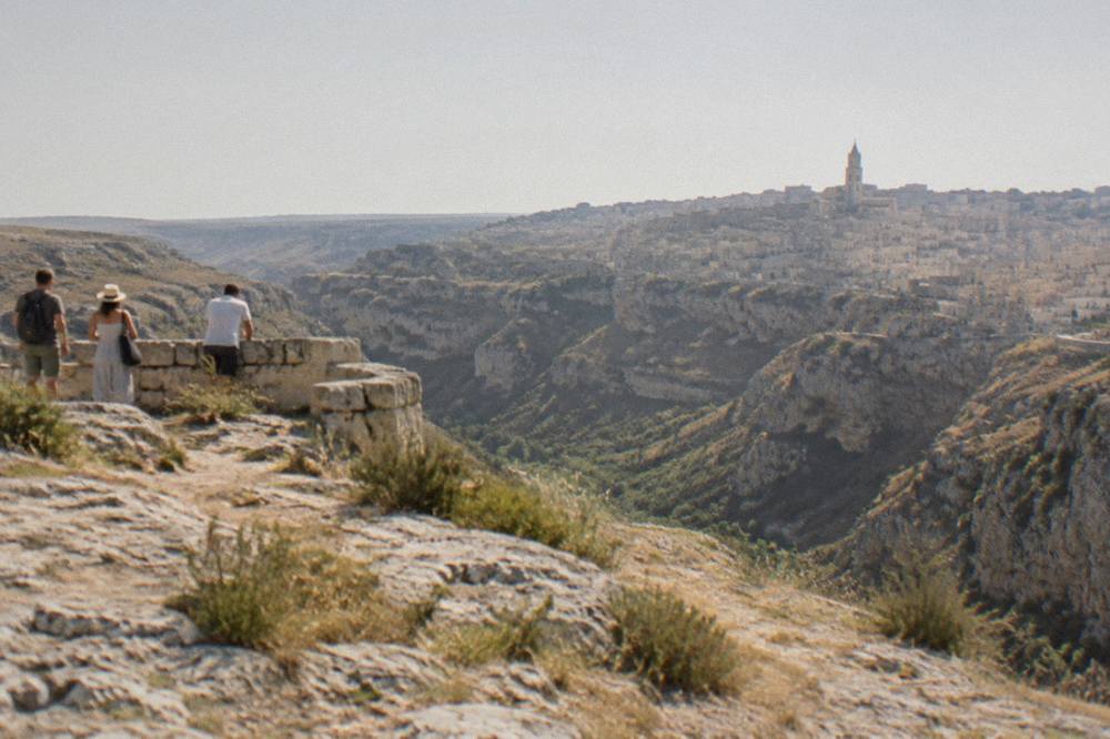 People standing at a stone viewpoint looking across the Gravina canyon toward the Sassi