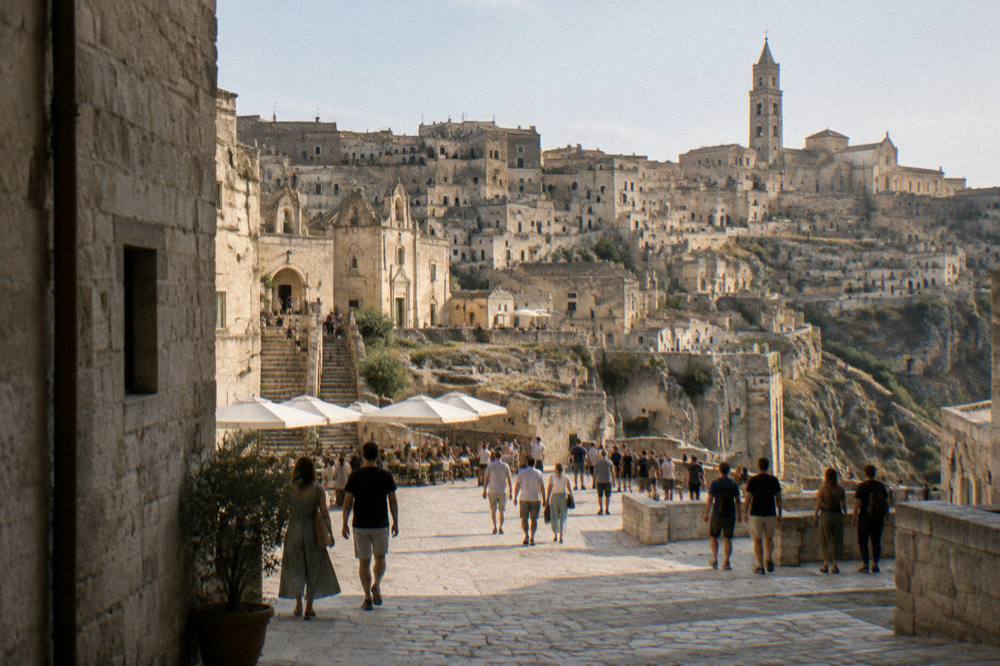 A crowded stone square in the Sassi of Matera on a warm evening with visitors walking