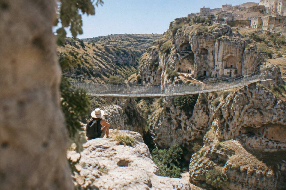 A hiker in a sun hat resting on a rock ledge above a canyon with cave churches carved in the cliff