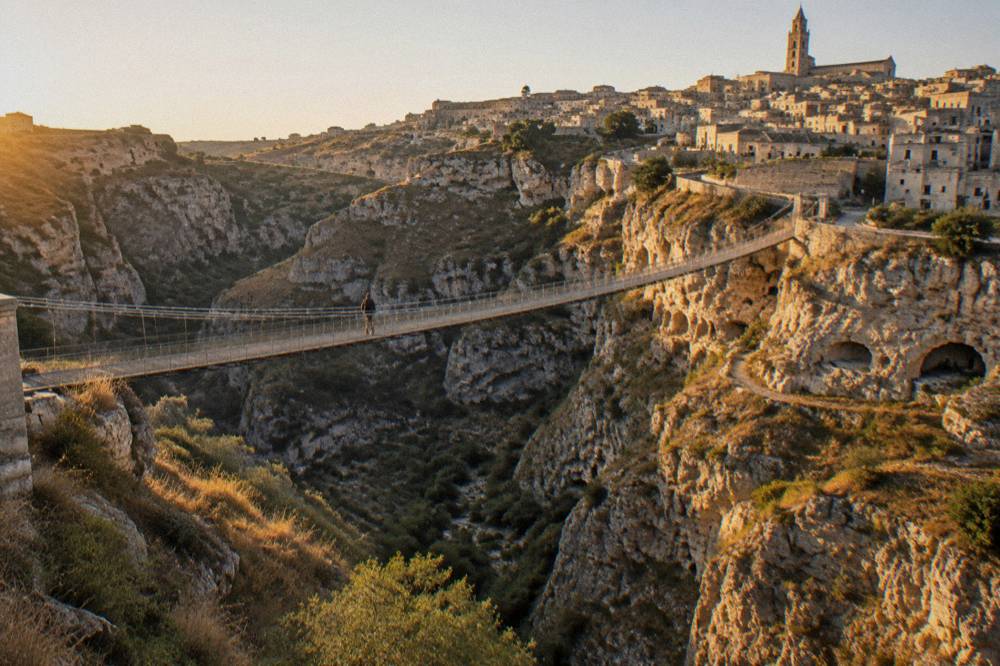 A wide dry canyon at golden hour with a stone city on the far rim and caves dotted along the cliff