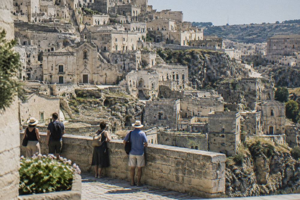 Visitors on a stone terrace overlooking the layered cave facades of the Sassi at midday