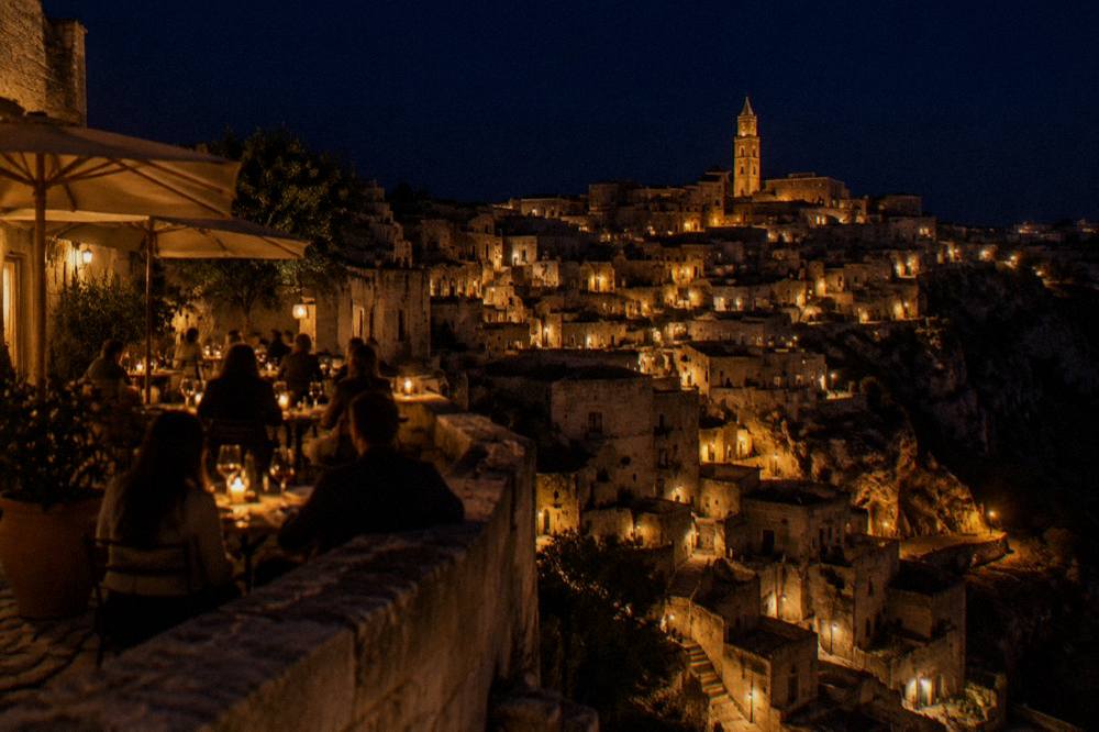 People dining at outdoor tables on a lit stone terrace above the Sassi at night