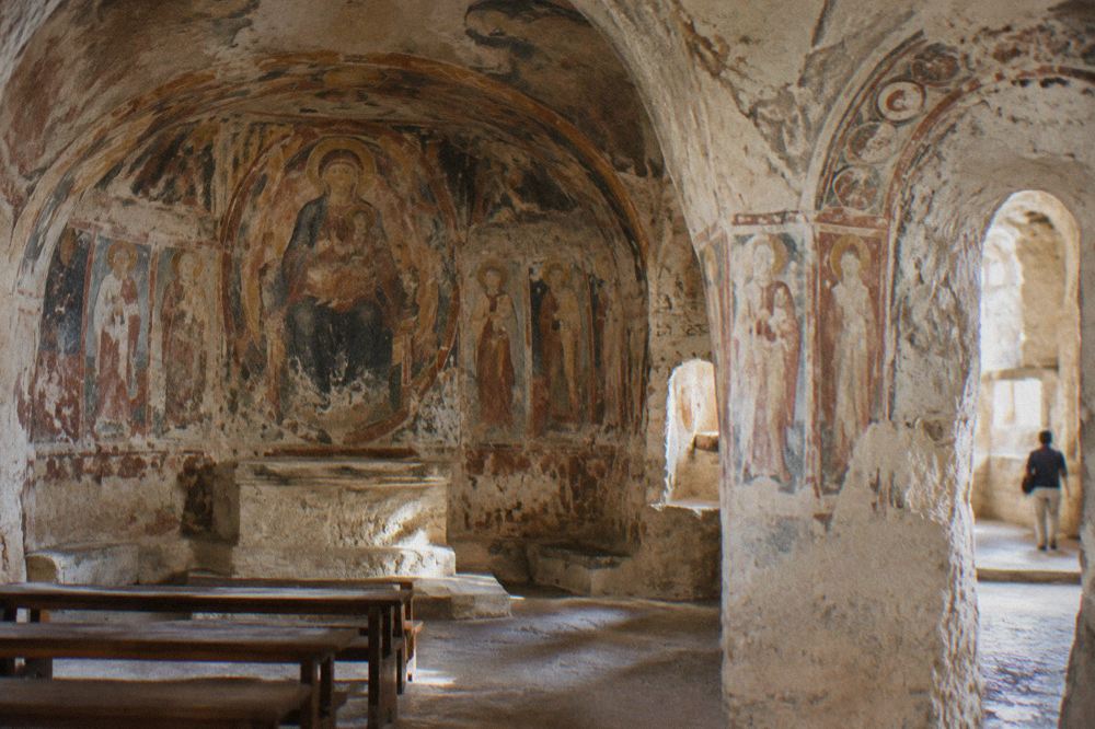 Interior of a cave church with frescoed walls lit softly in Matera