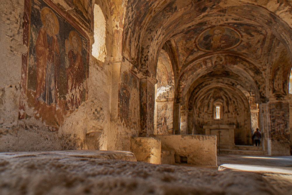 The interior of a rock-hewn church in Matera with frescoed walls lit softly and a barrel-vaulted ceiling